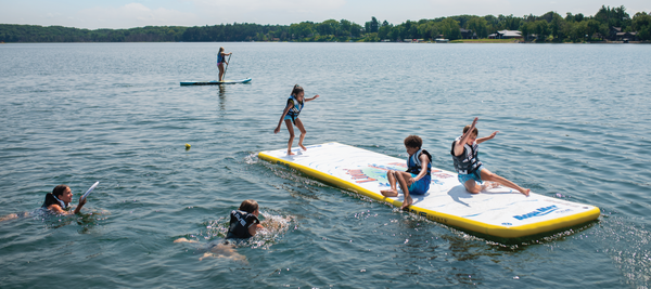 Children playing on a RAVE Sports Aqua Mat floating mat and another child on an inflatable stand up paddle board.
